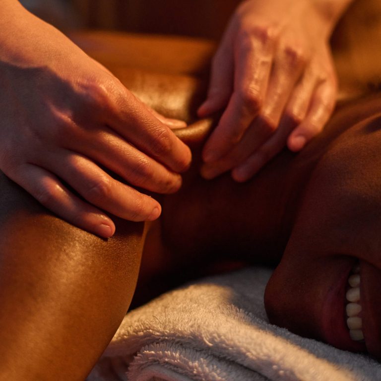 Medium close up of African American young woman squinting while unrecognizable masseuse using petrissage technique to massage deep shoulder muscles
